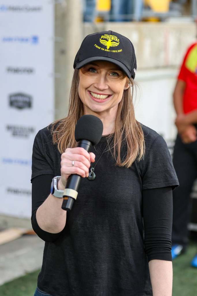 Kendall Forbes, wearing a Wellington Phoenix Football Club hat and holding a microphone at Sky Stadium to emcee Wrexham Down Under in Wellington